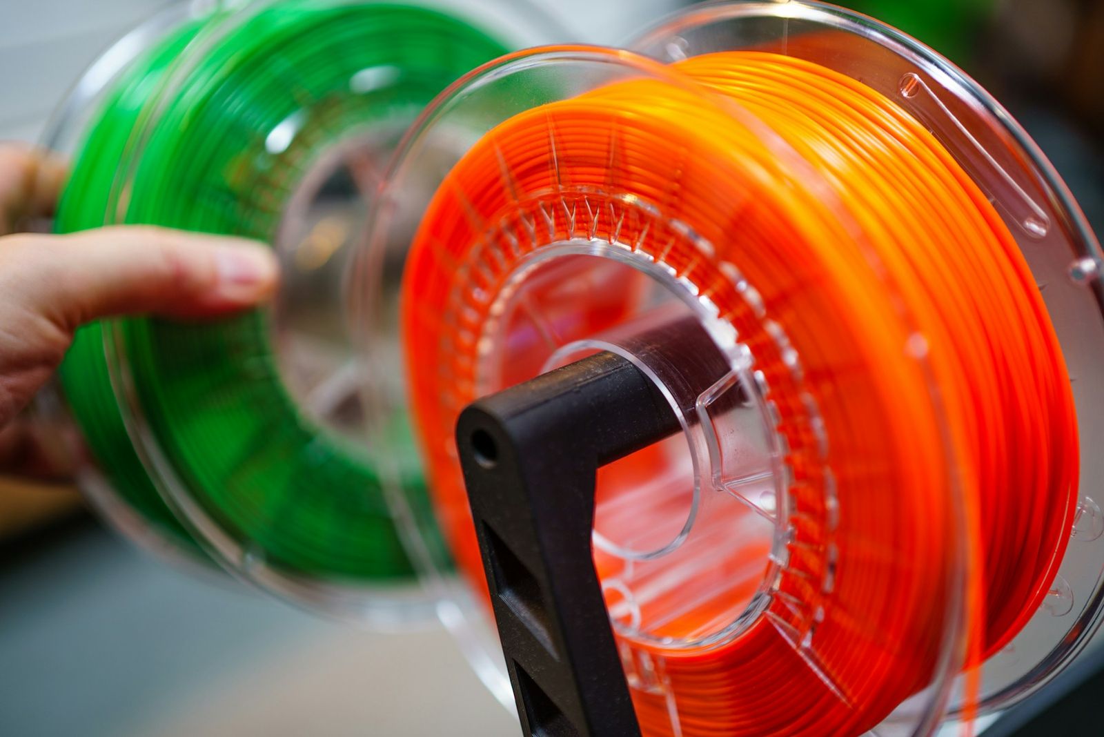 A hand holding green and orange filament spools on a spool holder